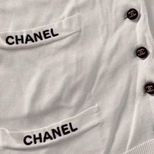 May include: White knitted cardigan with black embroidered "CHANEL" lettering on the pockets and gold buttons with the Chanel logo.
