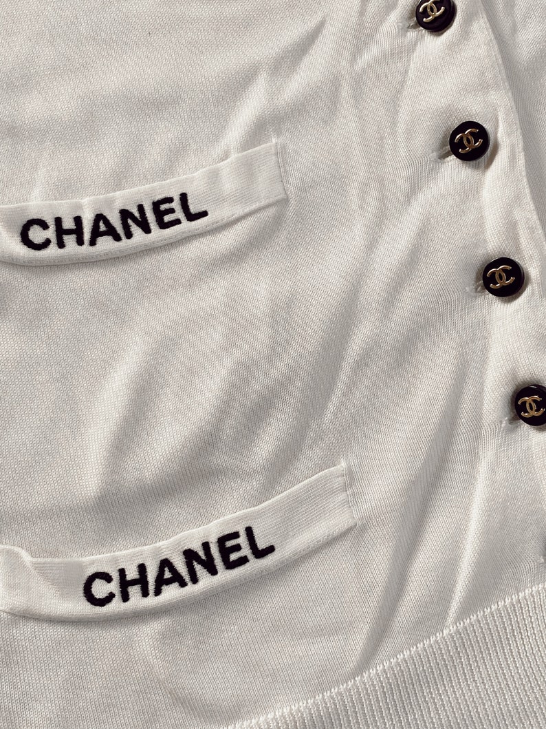 May include: White knitted cardigan with black embroidered "CHANEL" lettering on the pockets and gold buttons with the Chanel logo.