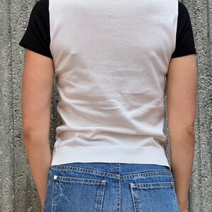 May include: A white knit shirt with black short sleeves. The shirt has a round neckline and a fitted silhouette. The model is wearing blue jeans.