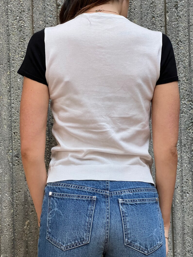 May include: A white knit shirt with black short sleeves. The shirt has a round neckline and a fitted silhouette. The model is wearing blue jeans.
