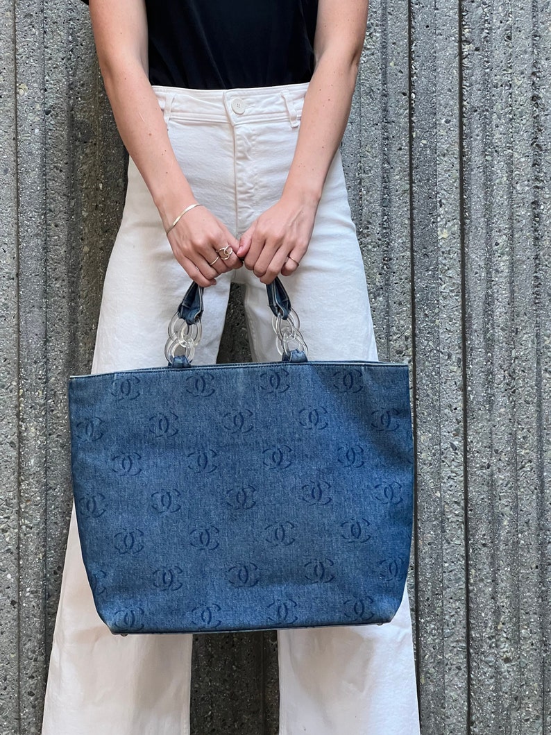 May include: A blue denim tote bag with a clear plastic handle and a repeating logo pattern. The bag is held by a person wearing white pants and a black shirt.