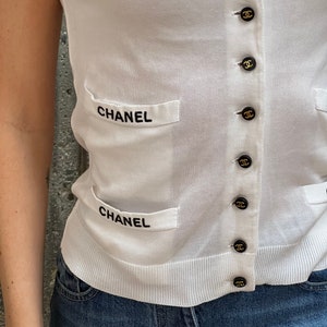 May include: White knitted cardigan with black trim and black buttons featuring the word "CHANEL" on the front pockets.