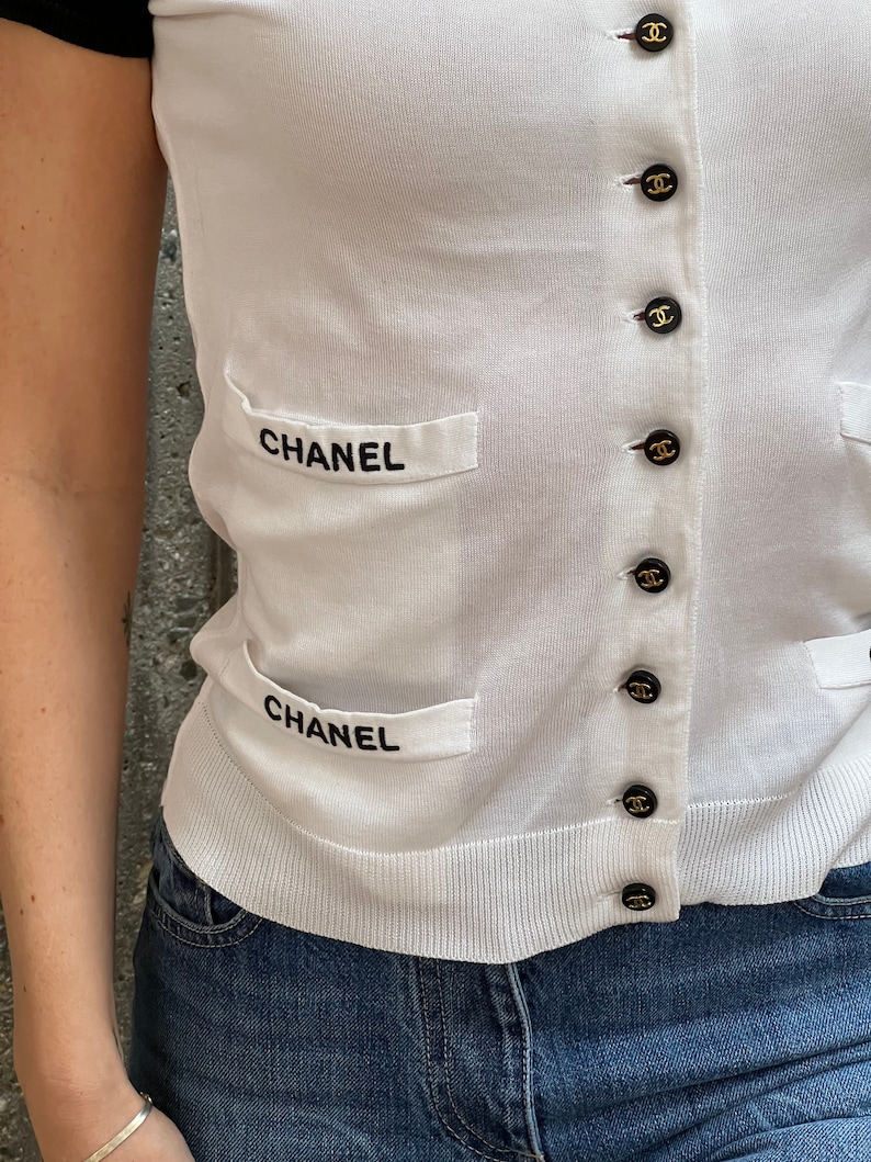 May include: White knitted cardigan with black trim and black buttons featuring the word "CHANEL" on the front pockets.