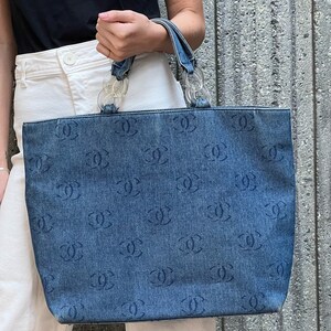 May include: A blue denim tote bag with a clear plastic chain handle. The bag is covered in a repeated pattern of the Chanel logo.