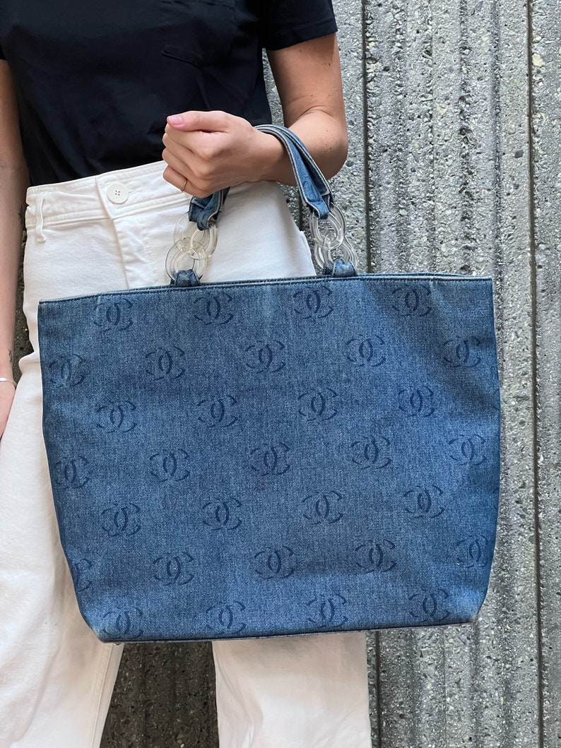 May include: A blue denim tote bag with a clear plastic chain handle. The bag is covered in a repeated pattern of the Chanel logo.