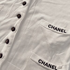 May include: White cardigan with black embroidered "CHANEL" on the pockets and black buttons with a gold Chanel logo.