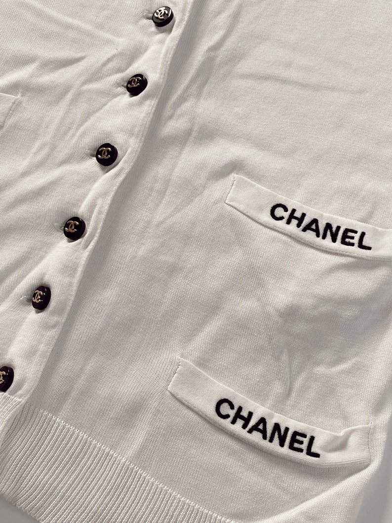 May include: White cardigan with black embroidered "CHANEL" on the pockets and black buttons with a gold Chanel logo.