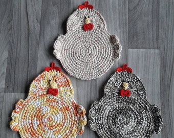 Pode incluir: Três bases para copos ou tachos em croché com formato de galinha. Uma é bege claro, uma é uma mistura variegada de amarelo, laranja e branco, e uma é uma mistura variegada de cinzento, preto e branco. Cada uma tem crista vermelha.