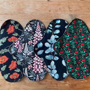 Puede incluir: Cuatro compresas de tela reutilizables con patrones distintos. La primera presenta amapolas naranjas sobre negro, la segunda flores de lupino rosa, la tercera hojas de eucalipto azul verdoso y la cuarta bayas rojas sobre follaje verde con una etiqueta negra.