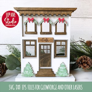 May include: A white wooden cutout of a cafe building with a brown roof and windows. The building has a sign that says "Cafe" and is decorated with green and red Christmas wreaths. The building is on a white base.