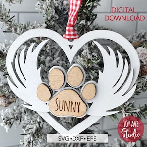 May include: White wooden heart-shaped ornament with a paw print in the center. The paw print is made of wood and has the name "SUNNY" engraved on it. The heart has wings on either side. The ornament is hanging on a Christmas tree.