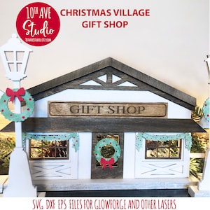 May include: A white wooden Christmas village gift shop with a brown roof and a sign that reads "Gift Shop". The shop has two windows with wreaths and garlands. The shop is lit up with string lights. SVG DXF EPS files for Glowforge and other lasers.