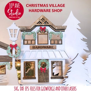 May include: A white wooden Christmas village hardware store with a brown roof and windows. The store has a sign that says "HARDWARE" and a wreath on the door. There are two white trees on either side of the store.