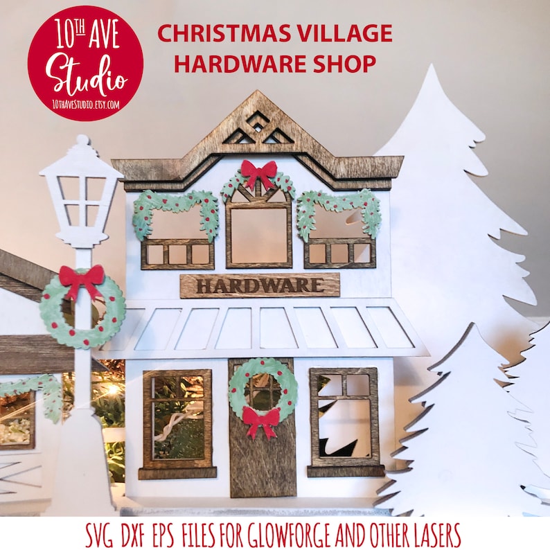 May include: A white wooden Christmas village hardware store with a brown roof and windows. The store has a sign that says "HARDWARE" and a wreath on the door. There are two white trees on either side of the store.