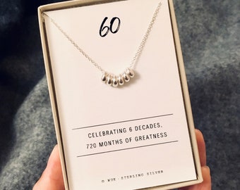 Può includere: Una scatola bianca con un ciondolo d'argento al suo interno. Il ciondolo ha 60 piccole perle d'argento. La scatola dice "Celebrating 6 decades, 720 months of greatness" e "KUE. Sterling Silver".