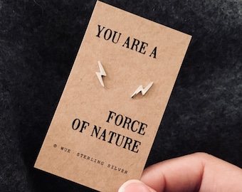 Puede incluir: Una tarjeta con el texto "You are a force of nature" y dos pendientes de plata en forma de rayo. La tarjeta está hecha de papel marrón y tiene el texto "WUE Sterling Silver" en la parte inferior.
