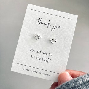 May include: Thank you for helping us tie the knot card with a pair of silver heart-shaped earrings. The card is white with black text. The earrings are on a white background.