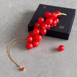 May include: A gold necklace with a red pendant and a cluster of red currant berries. The berries are next to a black box with the word "JULY" printed on it. A single red berry is also visible.