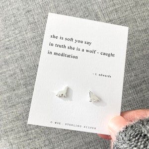 May include: A pair of sterling silver wolf stud earrings on a white card with the quote "she is soft you say in truth she is a wolf - caught in meditation" by t. edwards. The card is on a gray surface.
