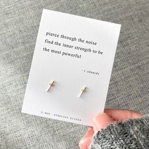 May include: A pair of sterling silver earrings featuring a small gold dagger design. The earrings are displayed on a white card with the text "pierce through the noise find the inner strength to be the most powerful - t. edwards" and "WUE STERLING SILVER".