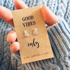 Könnte beinhalten: Nahaufnahme einer Karte mit dem Text "GOOD VIBES only" und einem kleinen silbernen Ananas-Anhänger auf jeder Seite. Die Karte wird von einer Hand gehalten, die einen grauen Pullover trägt. Die Karte enthält auch den Text "© WUE - STERLING SILVER".