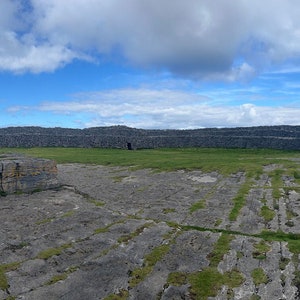 May include: A panoramic view of a rocky, grey landscape with a vast blue ocean in the background. The sky is a bright blue with white clouds.