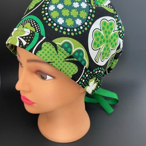 May include: Black scrub hat with a green and white shamrock pattern. The hat has a green tie.