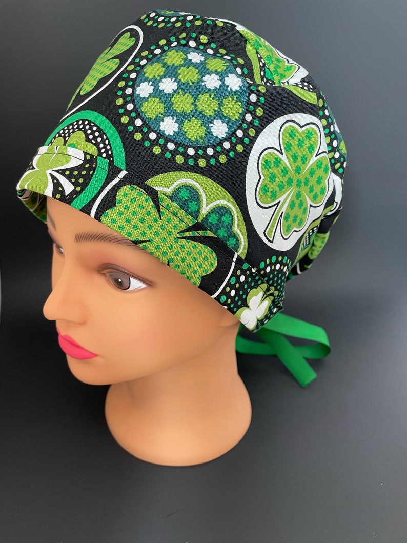 May include: Black scrub hat with a green and white shamrock pattern. The hat has a green tie.