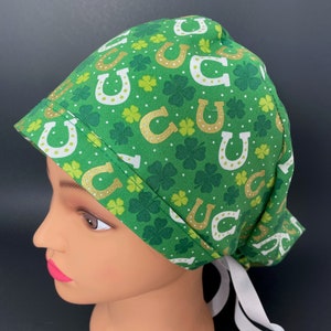 Puede incluir: Un gorro de quirófano verde con una cinta blanca. El gorro tiene un patrón de herraduras doradas y tréboles de cuatro hojas verdes.