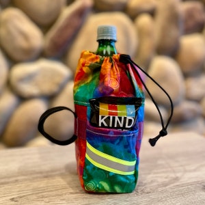Puede incluir: Un portabotellas colorido con cierre de cordón y un pequeño bolsillo que contiene una barra KIND. El soporte presenta un patrón de arcoíris con una franja reflectante y un lazo de transporte negro.