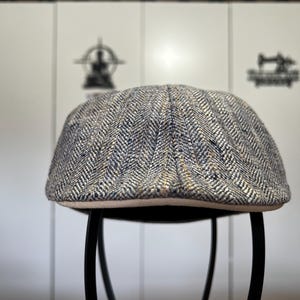 May include: A tweed newsboy cap with a herringbone pattern in shades of gray, blue, and tan. The cap has a rounded crown and a short, stiff brim. The cap is sitting on a black metal stool.