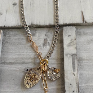 May include: A silver-toned curb chain necklace featuring a gold-toned connector and a dangling charm cluster. The charms include an oval metal tag embossed with a bee and the text QUEEN BEE, a hexagonal honeycomb charm with clear crystals and a small bee.