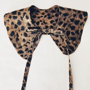 Pode incluir: Uma gola com estampa de leopardo com fecho de laço. A gola é marrom com manchas pretas e tem laços longos e finos. A gola é feita de um tecido macio e é provavelmente um acessório de moda.
