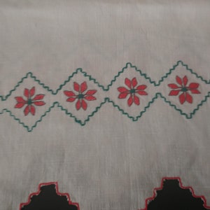 May include: A close-up of a white textile with a repeating pattern of red flowers inside green diamond shapes. The bottom edge features a black and red zigzag design. The fabric has a slightly wrinkled texture.