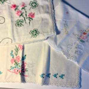 May include: Four white embroidered handkerchiefs with delicate floral designs. The handkerchiefs feature pink, blue, and green flowers and leaves. The handkerchiefs have a crocheted edge.