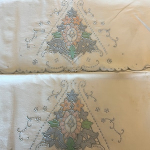 May include: Two white pillowcases with embroidered floral designs in shades of pink, green, and blue. The designs are in the shape of a triangle with a scalloped edge.