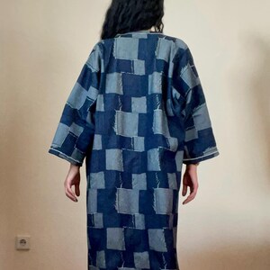 May include: A long, denim patchwork robe in shades of blue. The robe features a square pattern design and long sleeves. The person is facing away from the camera. The robe is knee-length and has a relaxed fit.