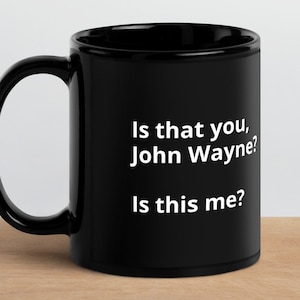 May include: A black ceramic mug with a white handle. The mug features the text "Is that you, John Wayne? Is this me?" in white lettering. The mug is sitting on a wooden surface.