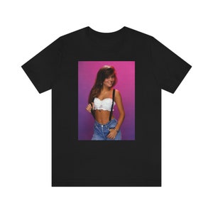 May include: A black t-shirt with a printed image of a woman wearing a white crop top and denim overalls. The woman has long brown hair and is standing in front of a pink and purple background.