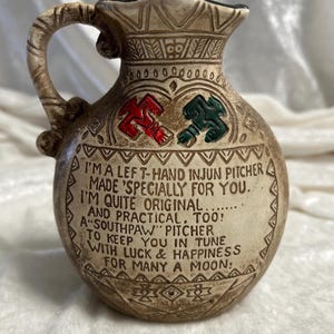 May include: A beige ceramic pitcher with a handle, featuring a red and green emblem and embossed text that reads: I'M A LEFT-HAND INJUN PITCHER MADE 'SPECIALLY FOR YOU. I'M QUITE ORIGINAL AND PRACTICAL, TOO! A "SOUTHPAW" PITCHER TO KEEP YOU IN TUNE WITH LUCK & HAPPINESS FOR MANY A MOON.