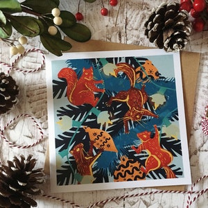 May include: Square greeting card with a playful illustration of squirrels set against blue and green foliage. The squirrels are in orange and brown hues. The card is on a brown envelope, with festive decorations.