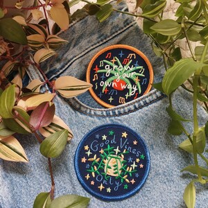 May include: Two embroidered patches on a blue denim background. One patch is blue with white stars and the text "Good Vines Only". The other patch is orange with white stars and the text "Grow your own way".