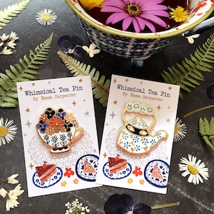 Puede incluir: Dos pines de esmalte con teteras caprichosas. El primer pin es una tetera con un diseño floral y el segundo pin es una tetera con un diseño floral azul y blanco y una máscara negra. Ambos pines están en tarjetas blancas con el texto "Whimsical Tea Pin By Emma Carpenter".