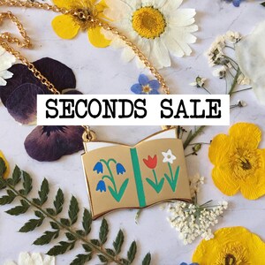 May include: A gold necklace with a book charm featuring colourful flowers. The charm is open to reveal a green and white interior. The text "SECONDS SALE" is displayed above the charm.