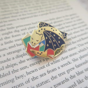 May include: A gold dragon enamel pin, featuring a navy wing design with star details. The dragon is depicted reading books with red, green, and blue covers. The pin is set on a background of open book pages.
