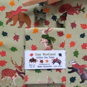 May include: A light green cotton tea towel with a woodland animal design, a rolling pin, and two metal cookie cutters. The towel features illustrations of a rabbit, bear, deer, and owl. The text "Cosy Woodland Cotton Tea Towel" is printed on the towel.