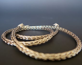 May include: Two silver chains, one braided and one woven, are intertwined and lying on a dark gray surface.