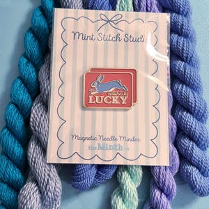 May include: A magnetic needle minder with a red and gold rectangular design featuring a blue rabbit and the words "naturally LUCKY." The needle minder is packaged with blue and purple yarn. The packaging reads "Mint Stitch Studio."