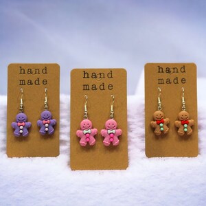 K&ouml;nnte beinhalten: Drei Paar handgemachte Lebkuchenmann-Ohrringe. Die Ohrringe befinden sich auf braunen Karten mit dem Text "hand made". Die Lebkuchenm&auml;nner sind lila, rosa und braun.