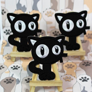 May include: Three black felt cat patches with white eyes and a black outline. The patches are displayed on small wooden easels against a background of paw print fabric.